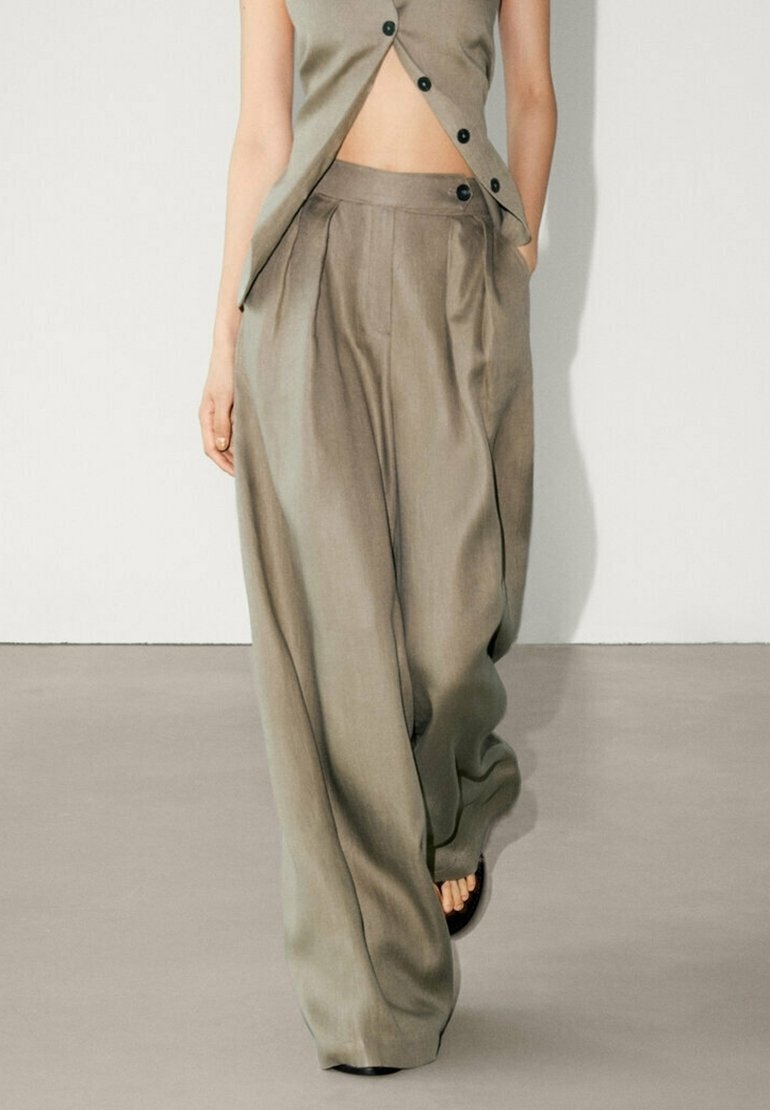 Femme portant un pantalon taupe taille haute, jambes larges avec plis, associé à un top sans manches boutonné, marchant sur un sol lisse.