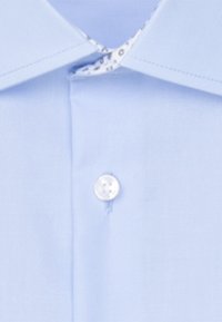 Jasnoniebieski kołnierzyk koszuli z białym guzikiem oraz częściowo widoczną wewnętrzną podszewką kołnierzyka z okrągłym wzorem.