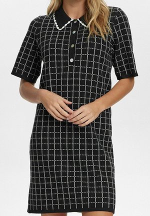 Femme portant une robe tricotée noire à manches courtes avec un motif à carreaux blancs, col festonné et patte de boutonnage à quatre boutons, mains lâchement jointes à la taille.