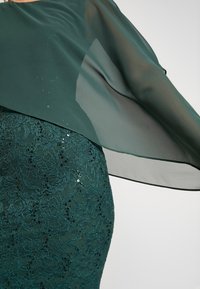 Robe en dentelle verte avec une superposition transparente. La dentelle présente des motifs floraux et des sequins. La superposition tombe élégamment des épaules.