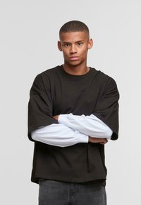 Sort oversized t-shirt med dropped shoulders lagd over en hvid langærmet skjorte. Rund hals, blødt bomuldsstof, afslappet pasform. Grå jeans.