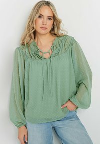 Blusa verde trasparente con pois in rilievo, scollatura arricciata e dettaglio con fiocco. Caratterizzata da maniche lunghe e una vestibilità comoda. Stile classico.