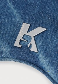 Sininen denim-kangas, jossa on kevyt rakenne. Hopeinen "K"-logo on näkyvästi esillä, ja siinä on monimutkaista yksityiskohtaisuutta ja brändäystä.