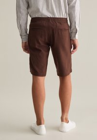 Bermuda marroni con vestibilità comoda, dotati di una vita con cordino e tasche posteriori. Abbinati a una camicia a righe a maniche lunghe e sneakers bianche.