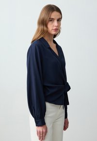 Blusa avvolgente blu navy con scollatura a V, maniche lunghe a palloncino e dettaglio annodato in vita. Realizzata in un tessuto morbido e leggero.