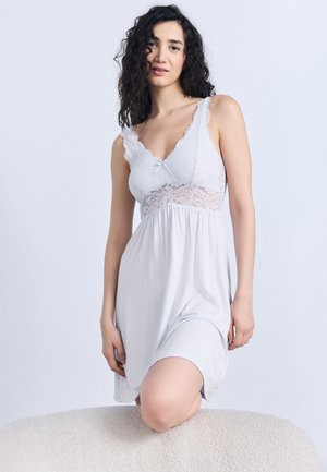 Vrouw met donker krullend haar, gekleed in een mouwloze witte kanten nachtjapon, knielend op een gevoerde ondergrond tegen een effen achtergrond