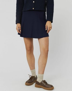 Femme portant une jupe marine, un pull bleu marine à texture et à boutons, des chaussettes beiges et des chaussures à lacets à semelle épaisse marron, se tenant devant un fond uni.