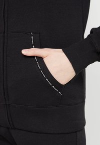 Sudadera con cremallera negra que presenta un bolsillo frontal con un borde del logotipo en blanco, hecha de una suave mezcla de algodón con puños de canalé.
