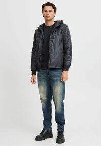Schwarze leichte Jacke mit Kapuze, Reißverschluss vorn und elastischen Bündchen, kombiniert mit hellblauen Jeans und schwarzen Schnürstiefeln.