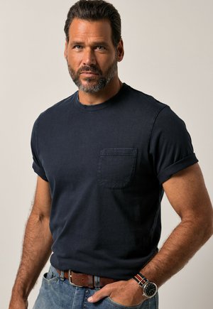 Marineblaues T-Shirt mit kurzen Ärmeln und rundem Halsausschnitt, ausgestattet mit einer Brusttasche, aus weichem Material gefertigt, getragen mit hellblauen Jeans und einem braunen Gürtel.