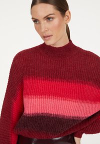 Frau mit heller Haut und braunen Haaren, die zurückgebunden sind, trägt einen rot-rosa gestreiften Rippstrickpullover mit einem Mock-Neck.