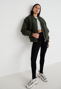 Olíva zöld bombázó dzseki, cropped szürke felső, fekete leggings és szürke sneaker. Simább textúrák, hangsúlyos zipzár és varrás részletekkel.