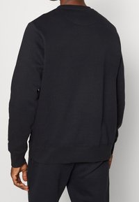 Uomo che indossa una felpa nera a maniche lunghe e pantaloni neri abbinati, visto da dietro su uno sfondo neutro.