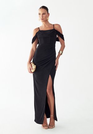 Vrouw draagt een zwarte off-shoulder jurk met een split aan de dij, houdt een gouden clutch vast, geaccentueerd met gouden oorbellen, armbanden en sandalen.
