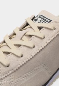 Κοντινή λήψη μπεζ σουέντ αθλητικού παπουτσιού Converse που δείχνει τα κρεμ κορδόνια δεμένα, λεπτομερή ραφή και μερικό λογότυπο της μάρκας στη γλώσσα.