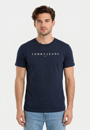 Giovane uomo con capelli castani corti indossa una t-shirt Tommy Jeans blu navy e jeans di denim chiaro, in piedi davanti a uno sfondo bianco uniforme.
