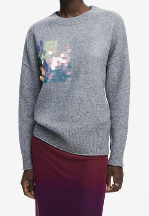Person iført en grå strikket sweater med iriserende pailletter på brystet og et nederdel med en gradient fra rød til lilla, hænder afslappet ved siden.