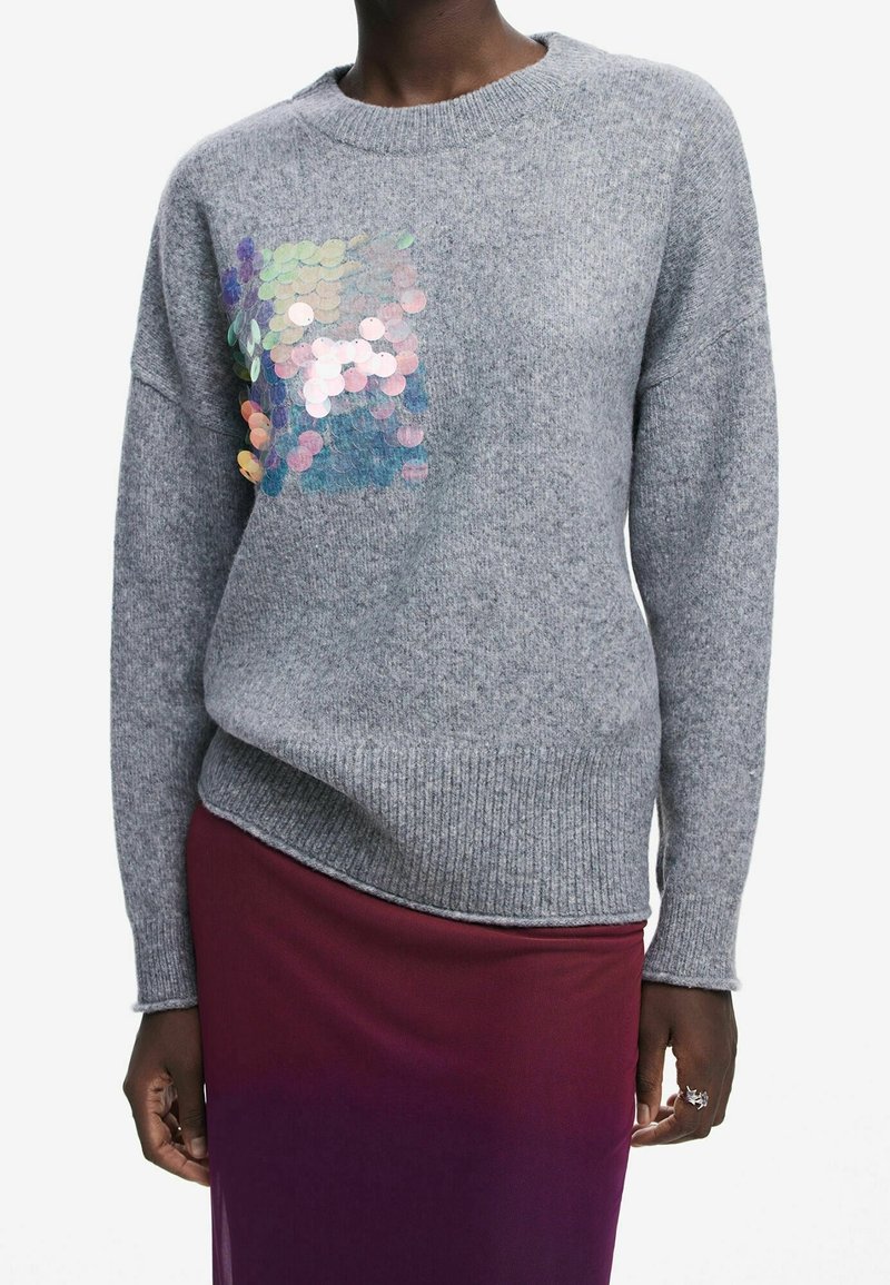 Personne portant un pull en maille gris avec des paillettes iridescentes sur la poitrine et une jupe dégradée du rouge au violet, mains détendues sur les côtés.