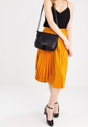 Bolso de hombro de cuero negro, falda midi amarilla plisada, blusa negra, zapatos de tacón negro con tiras en el tobillo. Texturas suaves, colores contrastantes.