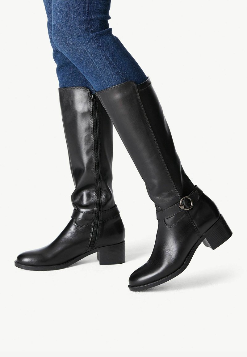 Jambes en jean bleu portant des bottes en cuir noires montant jusqu'aux genoux, avec des talons blocs bas et des fermetures éclair latérales, un pied légèrement levé.