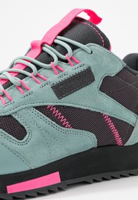 Sportovní obuv s modro-zeleným semišovým svrškem, černými síťovanými panely a růžovými akcenty. Mezi výrazné prvky patří zubatá podrážka a unikátní detaily šití.