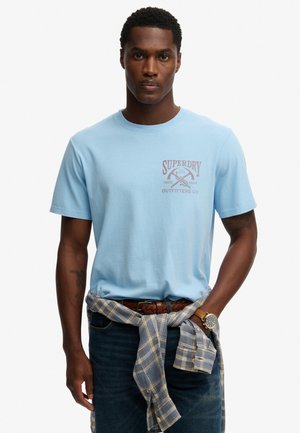 T-shirt en coton bleu clair à manches courtes, avec un imprimé graphique marron sur la poitrine. Porté avec une chemise à carreaux nouée à la taille et un jean en denim.