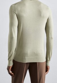 Světlounce zelený pletený svetr s dlouhými rukávy, žebrovanými lemy a hladkou texturou. Nošený přes hnědé kalhoty, ukazující střih na tělo.