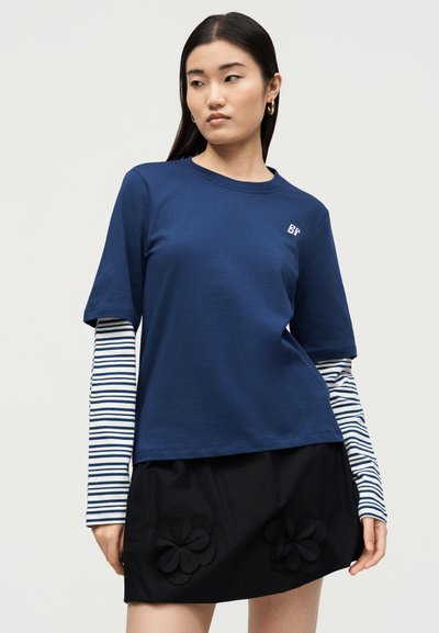 Νεαρή γυναίκα φοράει μπλε-σκούρο κοντομάνικο πουκάμισο πάνω από λευκή και μπλε-σκούρα ριγέ μακρυμάνικη μπλούζα και μαύρη φούστα με floral εφαρμογές.