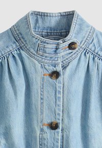 Jachetă din denim albastru deschis cu guler înalt, trei nasturi închiși vizibili și detalii de material adunat în apropierea umerilor.
