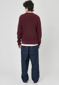 Pull en rib à manches longues de couleur bordeaux, col rond ; associé à un pantalon en denim bleu foncé à coupe décontractée avec poches arrière. Un t-shirt blanc est visible en dessous.