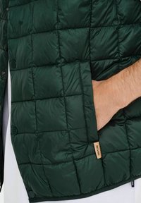 Gilet rembourré vert avec un motif matelassé, doté d'une poche latérale, de boutons-pression et d'une étiquette en tissu avec des lettres. Matériau lisse et brillant.