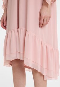 Rosa Chiffonkleid mit geschichteten, gefransten Saum. Leichter, luftiger Stoff zeigt eine sanfte Textur, knielang und locker geschnitten an den Ärmeln.