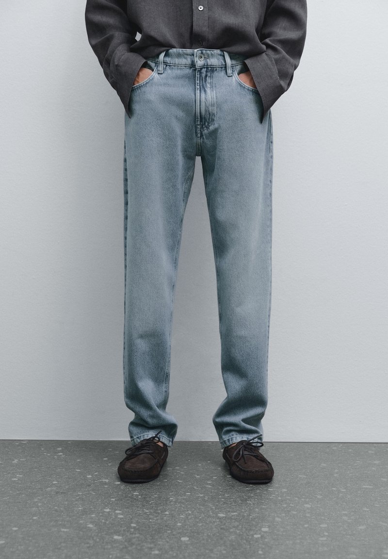 Personne portant un jean droit bleu clair, une chemise noire avec les manches baissées, les mains dans les poches et des chaussures à lacets brun foncé.