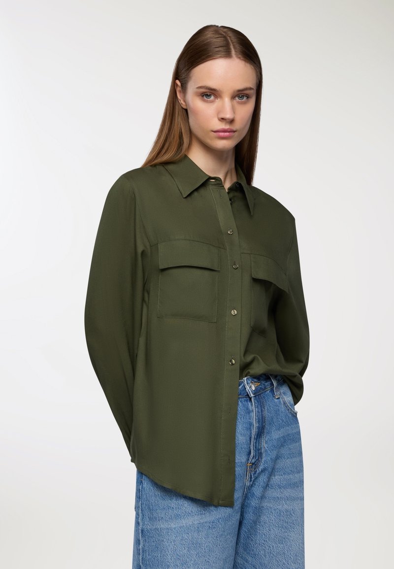 Blusa verde oliva con bottone sul davanti, due tasche sul petto e maniche lunghe. Realizzata in un tessuto leggero e liscio.
