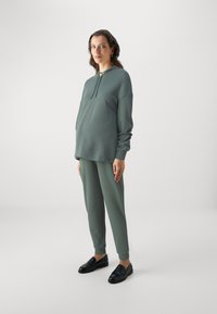 Green sweatshirt and joggers set in a soft fabric, featuring a hood and drawstring, paired with black shoes. Simple, relaxed fit design.