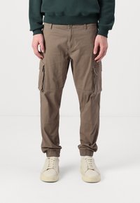 Man in bruin cargobroek met zijzakken en elastische boorden, gedragen met een donkergroene sweatshirt en crème kleurige sneakers.