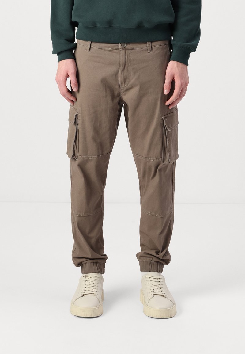 Man in bruin cargobroek met zijzakken en elastische boorden, gedragen met een donkergroene sweatshirt en crème kleurige sneakers.