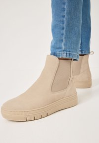 Bottines en suède beige avec des inserts latéraux en �élastique et des semelles en caoutchouc texturé, associées à un jean en denim bleu.