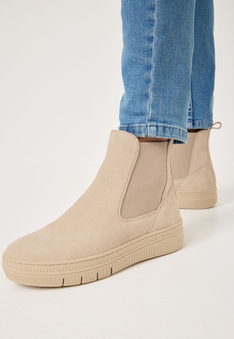 Bottines en suède beige avec des inserts latéraux en élastique et des semelles en caoutchouc texturé, associées à un jean en denim bleu.