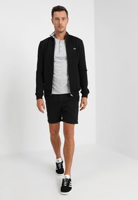 Schwarze Zip-Jacke über einem grauen Henley-Shirt, schwarze Shorts und schwarze Sneakers mit weißen Akzenten. Lässiges sportliches Outfit.