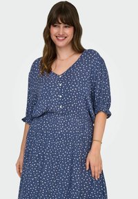 Zila rakstu kleita ar v-veida kakla izgriezumu, īsām piedurknēm un pogu detalizāciju. Iezīmēta ar pieguļošu vidukli un plūstošu svārku ar baltām ziedu nospiedumiem.