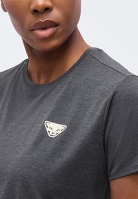 Κοντινό πλάνο ενός ατόμου που φοράει σκούρο γκρι μπλουζάκι με λευκό τριγωνικό λογότυπο προσώπου πάνθηρα στο στήθος.