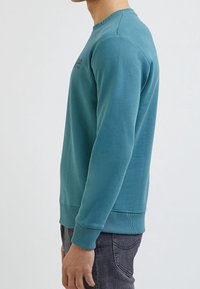 Modrá mikina s dlouhými rukávy, kulatým výstřihem a žebrovanými manžetami. Na hrudi je jemné logo a materiál je hladký bavlněný.