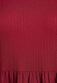 Tejido rojo texturizado con una superficie arrugada. Presenta un dobladillo con volantes en la parte inferior. El diseño general es simple y monocromático.