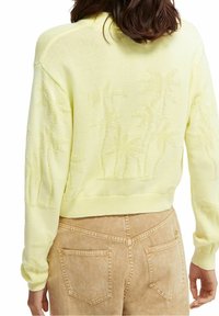Suéter amarillo claro con diseño texturizado de palmera, puños y dobladillo acanalados, combinado con pantalones marrón claro que tienen bolsillos traseros.