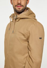 Lichtbruine jas met capuchon, voorzien van een voorrits en trekkoorden, gestructureerde stof en een klein logopatch op de mouw.