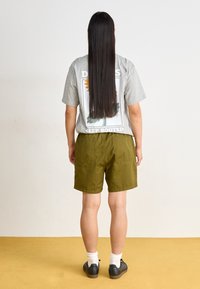 Sivý oversized tričko s grafickým potlačou, zelené šortky, biele ponožky a čierne tenisky. Jednoduché pozadie, žltá podlaha.