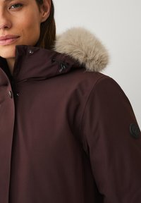 Bordeauxrode buitenjas met een rand van nepbont aan de capuchon, verstelbare trekkoorden en een zwarte embleem op de mouw. Strakke, gestructureerde stof.