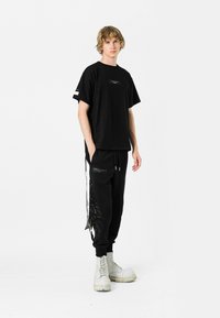 Camiseta negra de gran tamaño con un bolsillo en el pecho; combinada con pantalones jogger negros con bolsillos con cremallera y un diseño de plumas en el costado. Botas blancas.