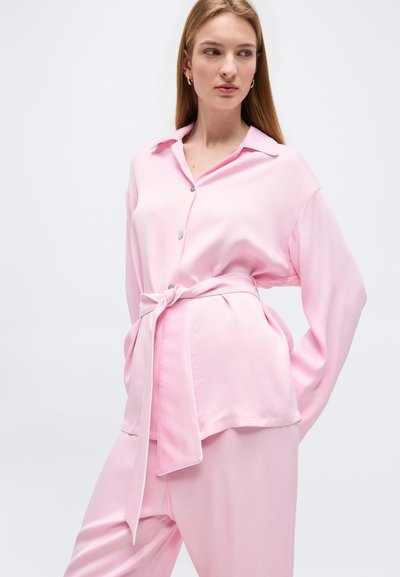 Γυναίκα που φοράει ανοιχτό ροζ σατέν μπλούζα με δεμένη ζώνη και παντελόνι σε ίδιο χρώμα, στέκεται με τα χέρια στις τσέπες μπροστά σε απλό φόντο.